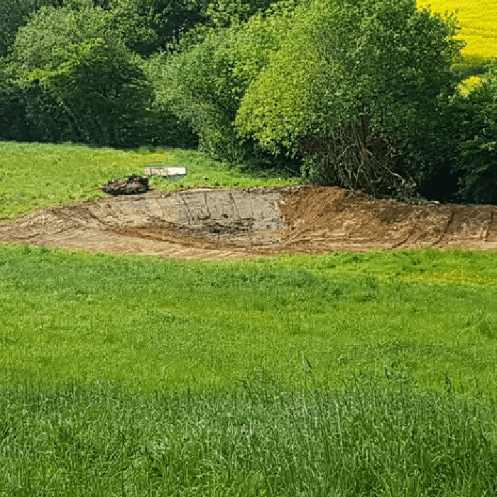 Image of a pond under construction. We see a large hole carved out of a field next to a stream.